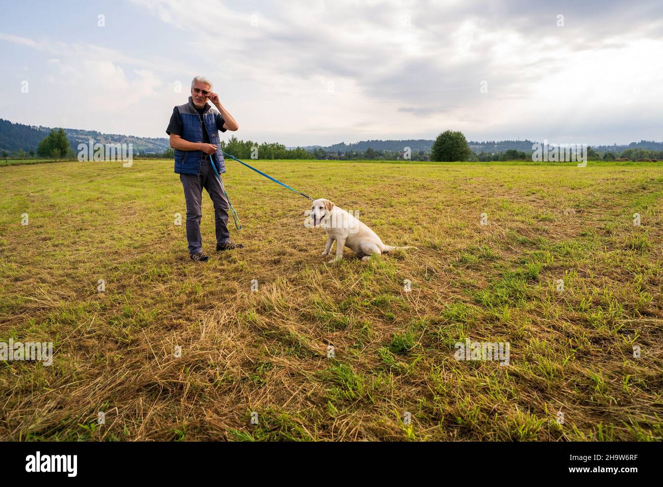 Zakopane, Polen - 15. August 2021: Mann steht in einem grünen Feld auf dem Land und hält eine Schnur, die an einen goldenen labrador-Retriever-Hund gebunden ist Stockfoto