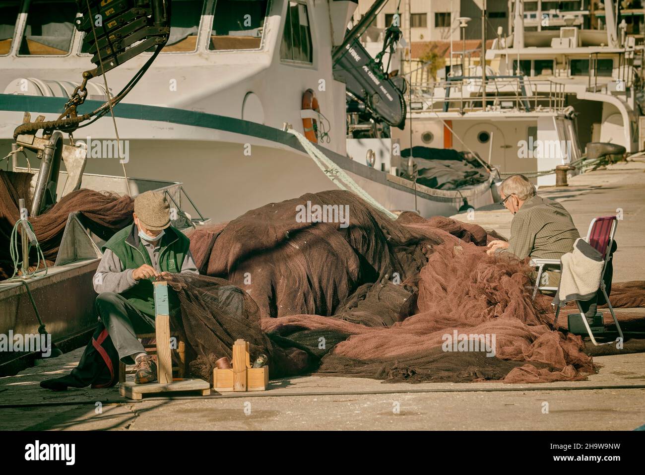 Zwei Männer, die auf dem Pier des Hafens der Stadt Burriana mit den Fischerbooten im Hintergrund Fischernetze nähen, Castellon, Spanien Stockfoto