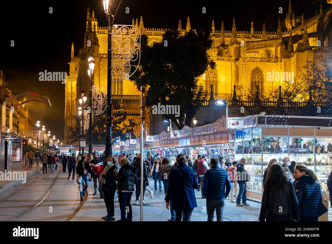 Nacht auf dem Weihnachtsmarkt, während Touristen und einheimische Spanier die Dekorationen mit der Kathedrale von Sevilla im Blick hinter sich genießen. Stockfoto