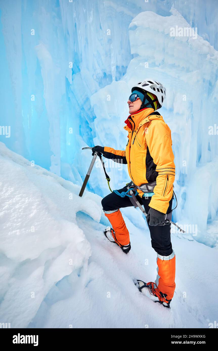 Klettererin in der Nähe von gefrorenem Wasserfall im Helm mit Eispickel in orangefarbener Jacke in den Bergen. Sportbergsteigen und Alpinismus Konzept. Stockfoto