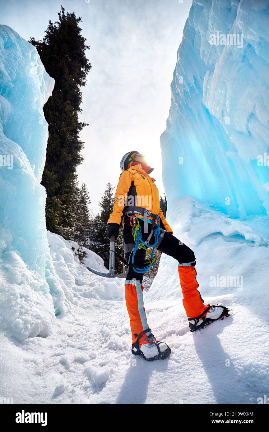 Klettererin in der Nähe von gefrorenem Wasserfall im Helm mit Eispickel in orangefarbener Jacke in den Bergen. Sportbergsteigen und Alpinismus Konzept. Stockfoto