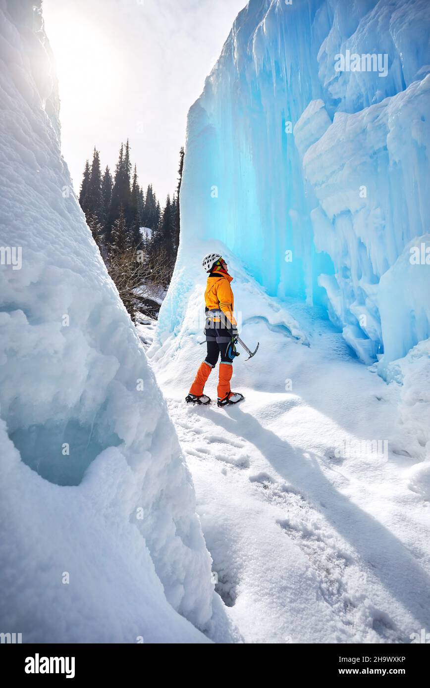 Klettererin in der Nähe von gefrorenem Wasserfall im Helm mit Eispickel in orangefarbener Jacke in den Bergen. Sportbergsteigen und Alpinismus Konzept Stockfoto