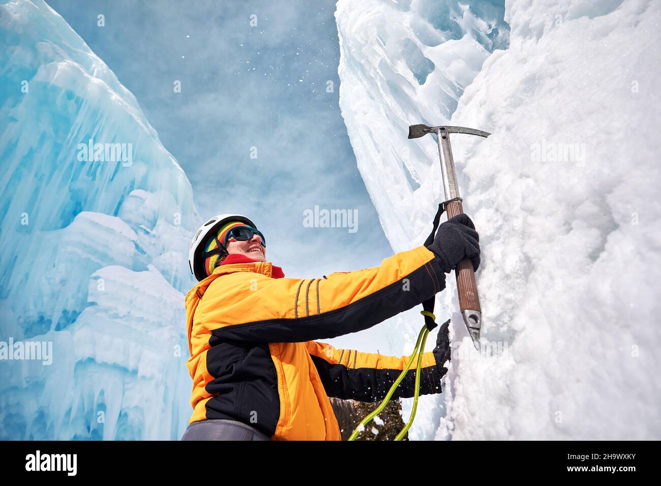 Die Frau klettert in den Bergen einen gefrorenen Wasserfall im Helm mit Eispickel in orangefarbener Jacke. Sportbergsteigen und Alpinismus Konzept. Stockfoto