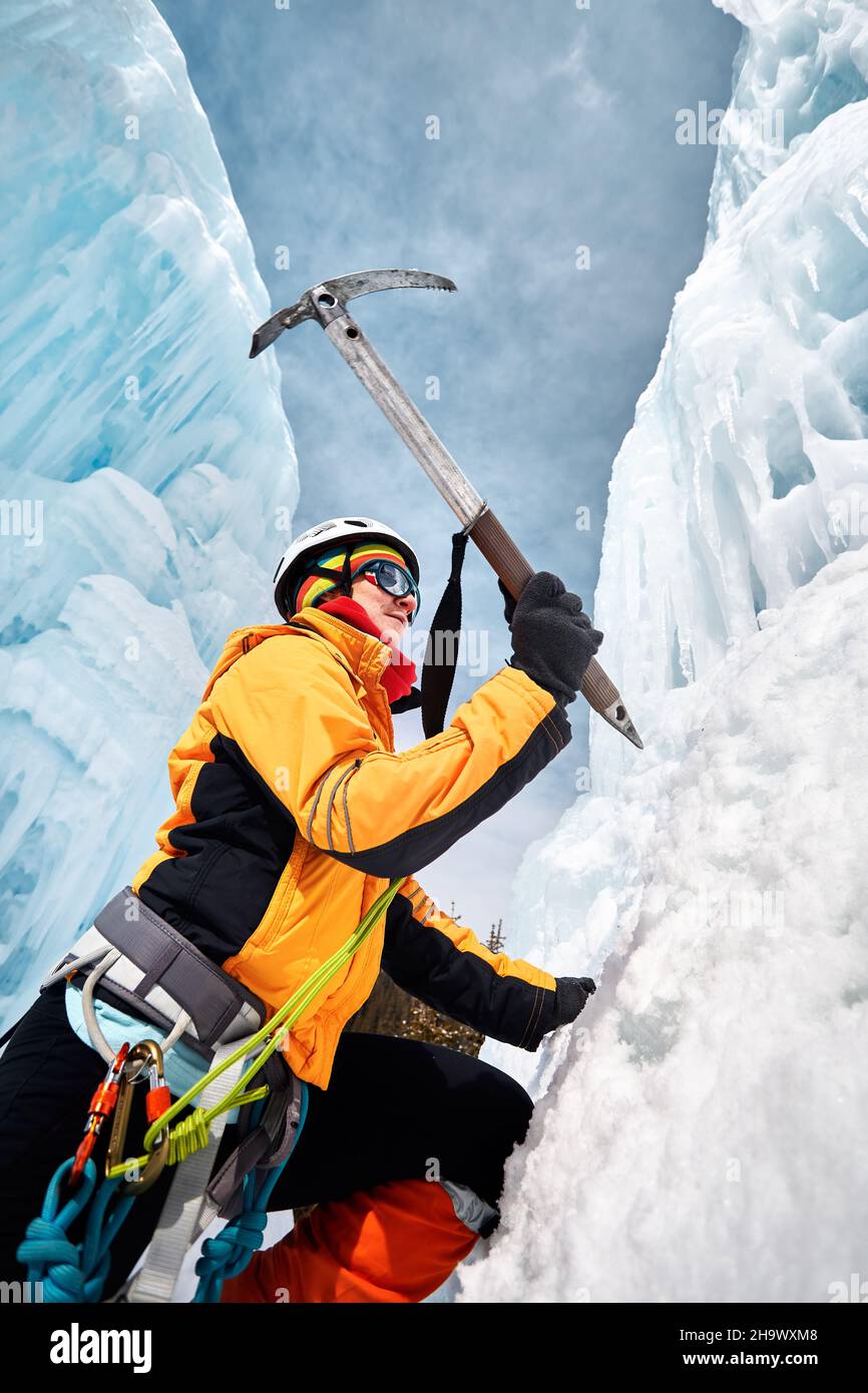 Die Frau klettert in den Bergen mit Eispickel in orangefarbener Jacke über einen gefrorenen Wasserfall. Sportbergsteigen und Alpinismus Konzept. Stockfoto