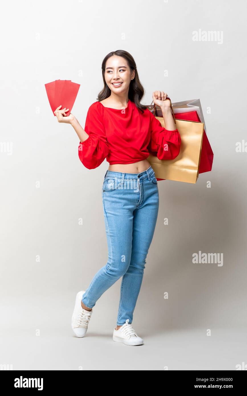 Lächelnde asiatische Frau hält Einkaufstaschen und rote Umschläge oder Ang Pao in isolierten hellgrauen Studio Hintergrund für Lunar Neujahr Verkaufskonzept Stockfoto