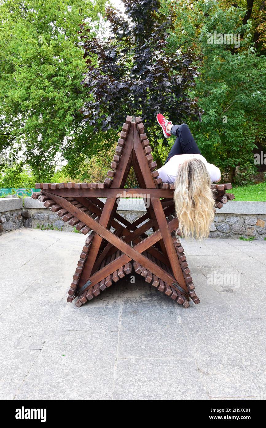 Auf einer dekorativen Holzbank in Form eines 5-spitzen Sterns liegt ein blondes Mädchen mit langen Haaren auf dem Rücken Stockfoto