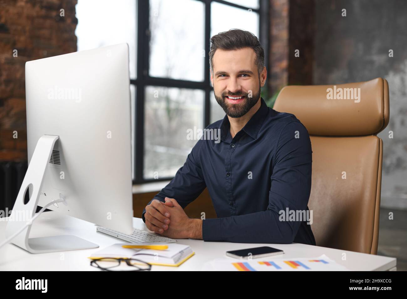 Optimistischer, männlicher Büroangestellter, der am Schreibtisch vor dem PC sitzt, die Kamera anschaut und lächelt, erfolgreicher junger Geschäftsmann im modernen Loft-Büro, Manager-ceo in eleganter Casual-Atmosphäre am Arbeitsplatz Stockfoto