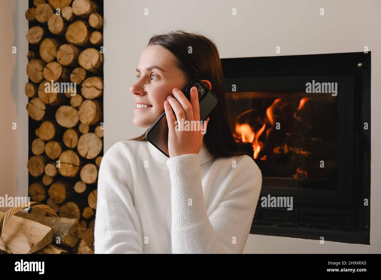 Lächelnde junge kaukasische Frau in weißem Pullover, die auf warmem Boden in der Nähe des Kamins zu Hause sitzt und sich mit dem Handy unterhält Stockfoto