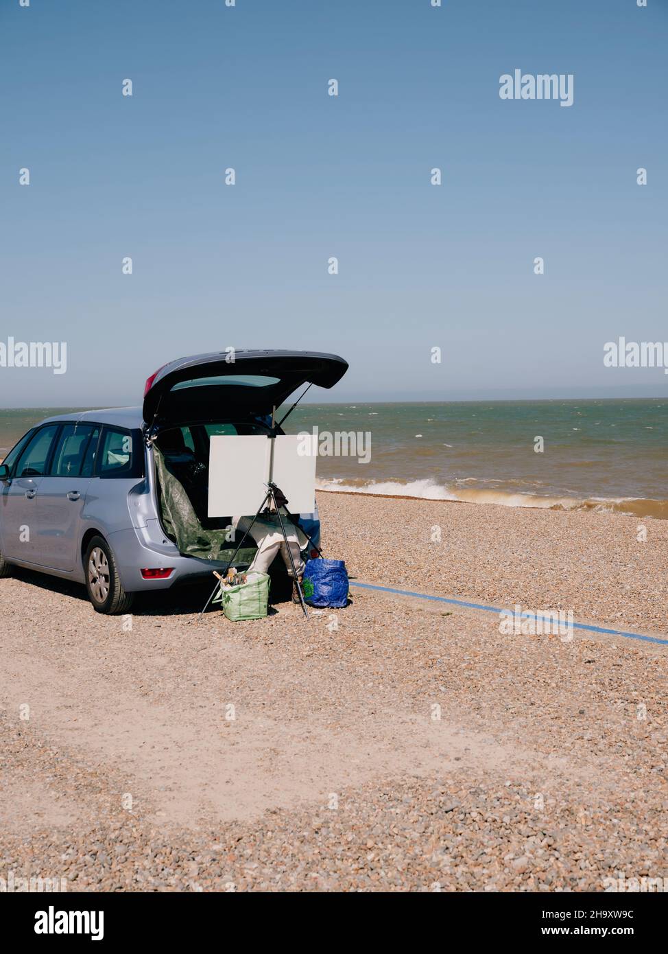 Ein Künstler malt aus dem Kofferraum seines Autos mit einer Staffelei und Leinwand an der Sommerküste Großbritanniens - Plein air painting - en plein air Stockfoto