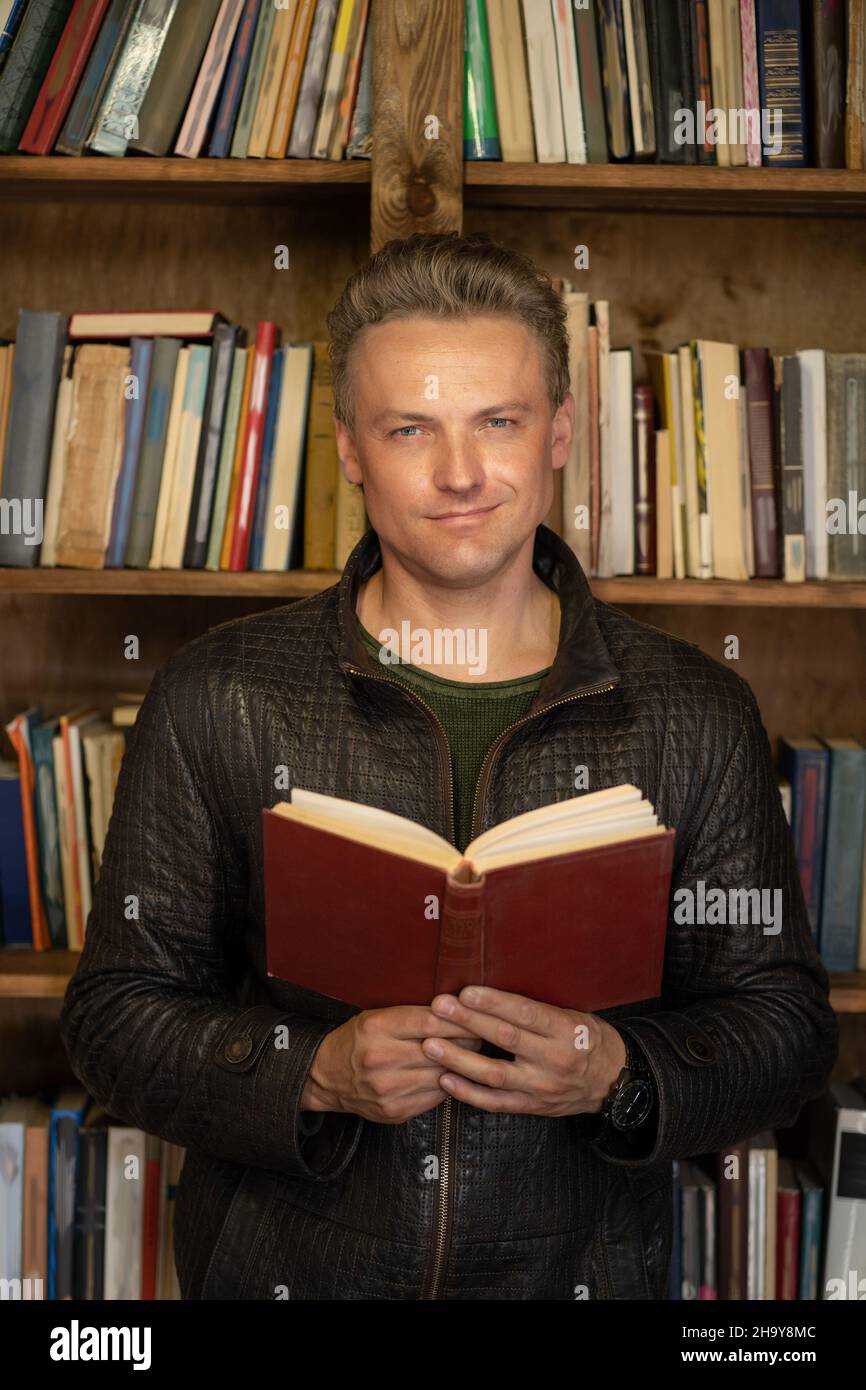 Selbstbewusster, hübscher Mann mit einem roten Vintage-Buch in den Händen steht vor dem Hintergrund von Regalen mit Büchern der Privatbibliothek. Nahaufnahme Porträt. Hochwertige Fotos Stockfoto