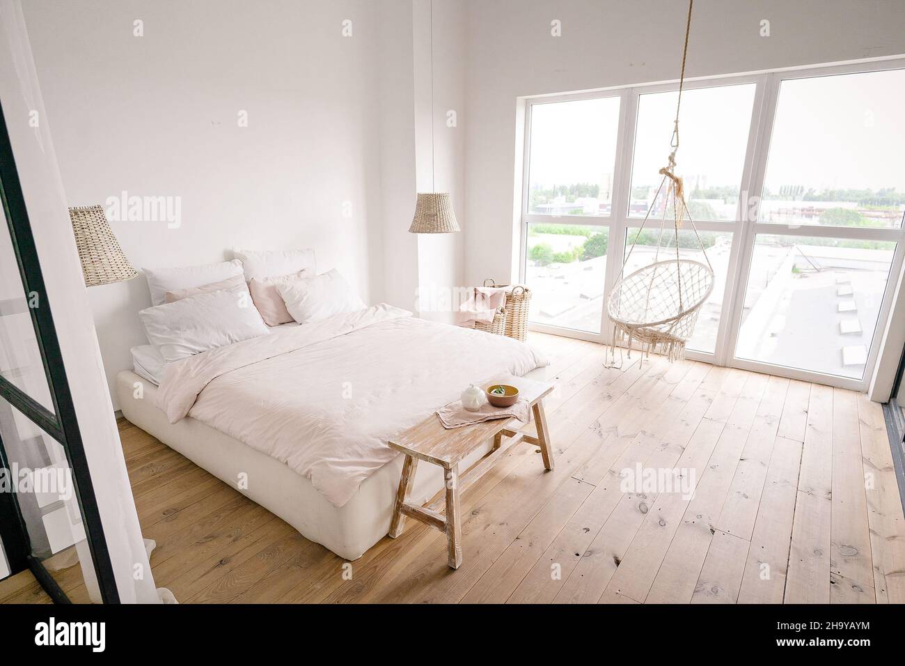 Großes helles Schlafzimmer in minimalistischem Stil in Pastelltönen mit Doppelbett, weißen Wänden, einem Wicker-Wäschekorb, Korbleuchtern, beigefarbener Bettbank, großem Fenster, Boho-Hängestuhl und Holzboden. . Hochwertige Fotos Stockfoto