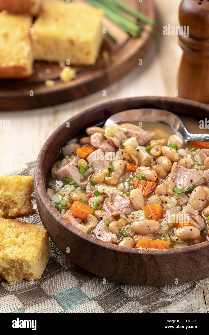 Schinken- und Bohnensuppe mit Karotten und Schnittlauch in einer Holzschüssel mit Maisbrot auf einem rustikalen Holztisch Stockfoto