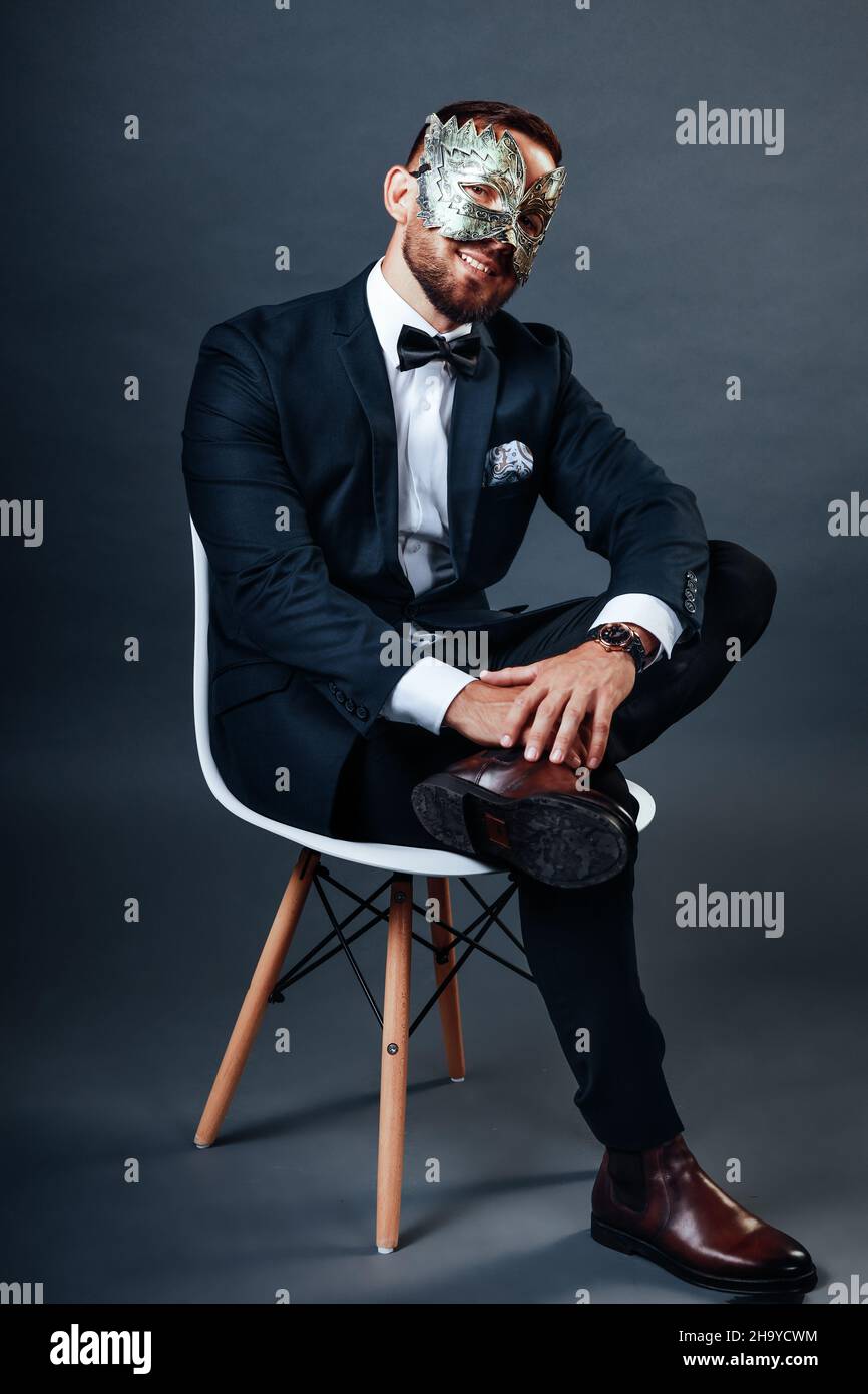Der Mann im Business-Anzug sitzt auf dem Stuhl und trägt eine Karnevalsmaske auf seinem Gesicht. Studio-Porträt auf grauem Hintergrund. Ein junger Mann von 25-30 Jahren versteckt sein Gesicht unter Maske. Listiger und hinterlistiger Geschäftsmann. Trickster. Stockfoto