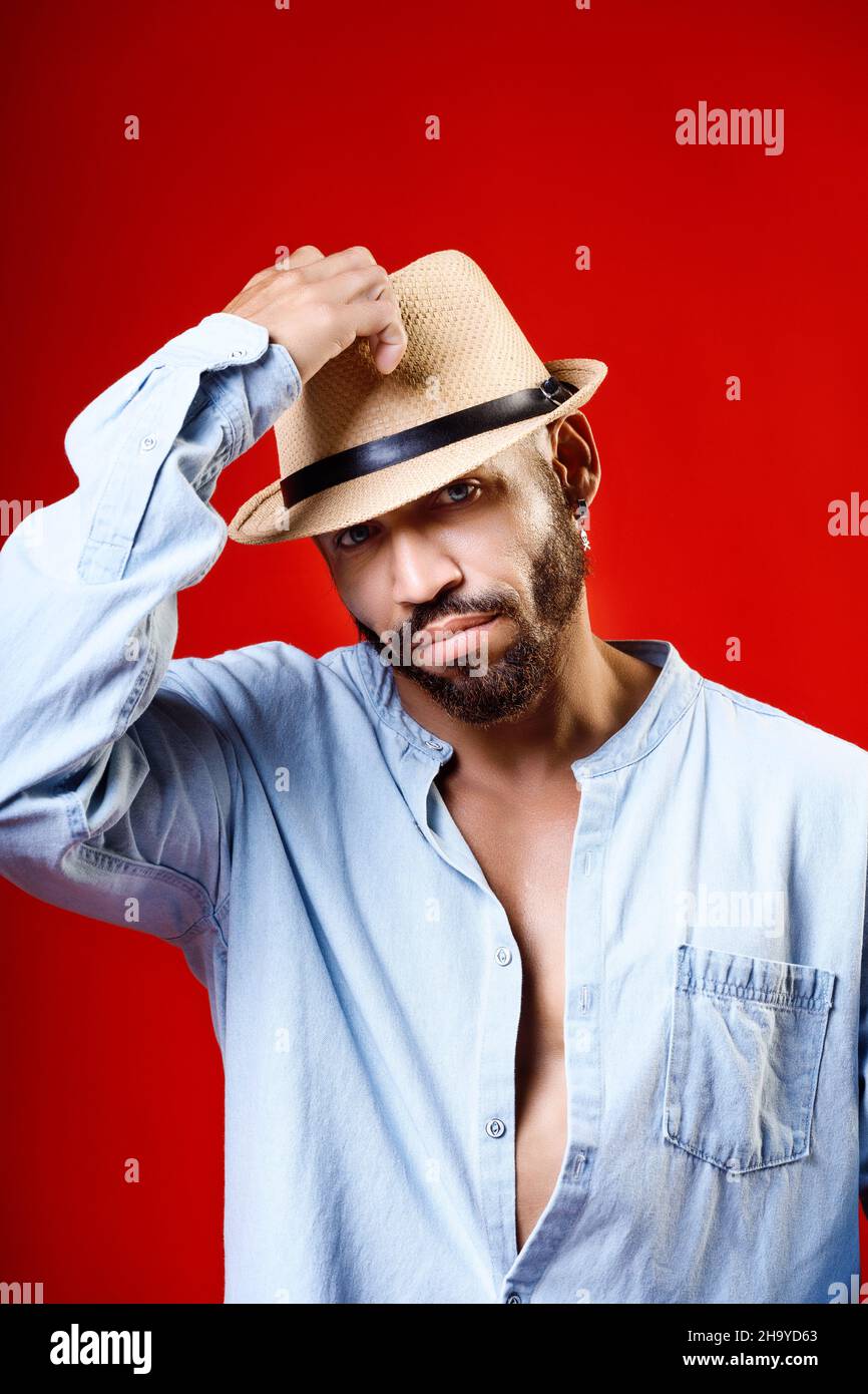 Kubanischer oder lateinamerikanischer Mann in Hut und Hemd auf rotem Hintergrund. Studioportrait des bärtigen Mannes 30-40 Jahre alt. Neutrale Emotionen. Der Mann nimmt seinen Hut vom Kopf. Stockfoto