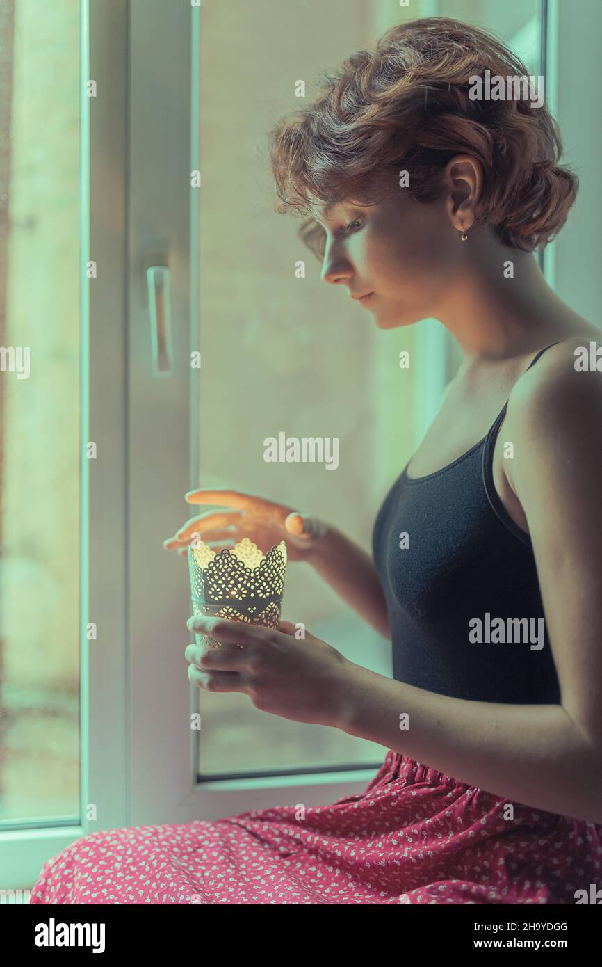 Silhouette einer schönen jungen Dame mit braunem lockigen Haar in authentischer Kleidung, die auf dem Fensterbrett sitzt. Die Hände des Mädchens halten einen Kerzenständer Stockfoto