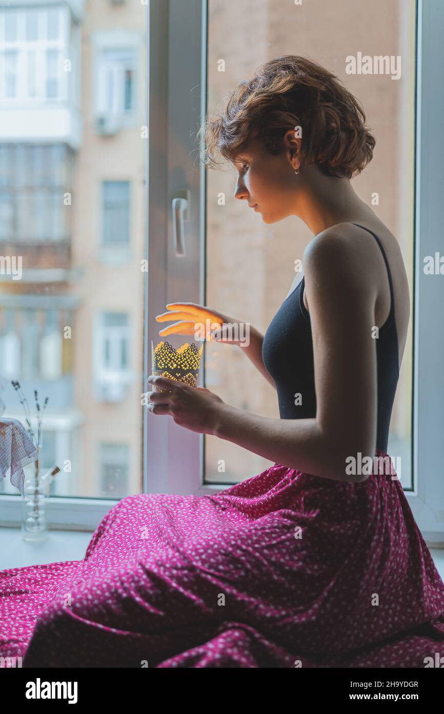 Silhouette einer schönen jungen Dame mit braunem lockigen Haar in authentischer Kleidung, die auf dem Fensterbrett sitzt. Die Hände des Mädchens halten einen Kerzenständer Stockfoto