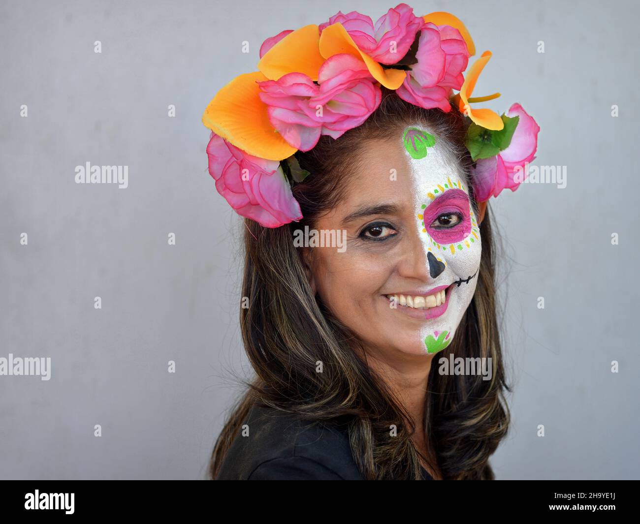 Mexikanische Frau mit bunten Blumen im Haar malt die linke Hälfte ihres Gesichts mit Catrina Face Make-up am Tag der Toten und lächelt den Betrachter an. Stockfoto