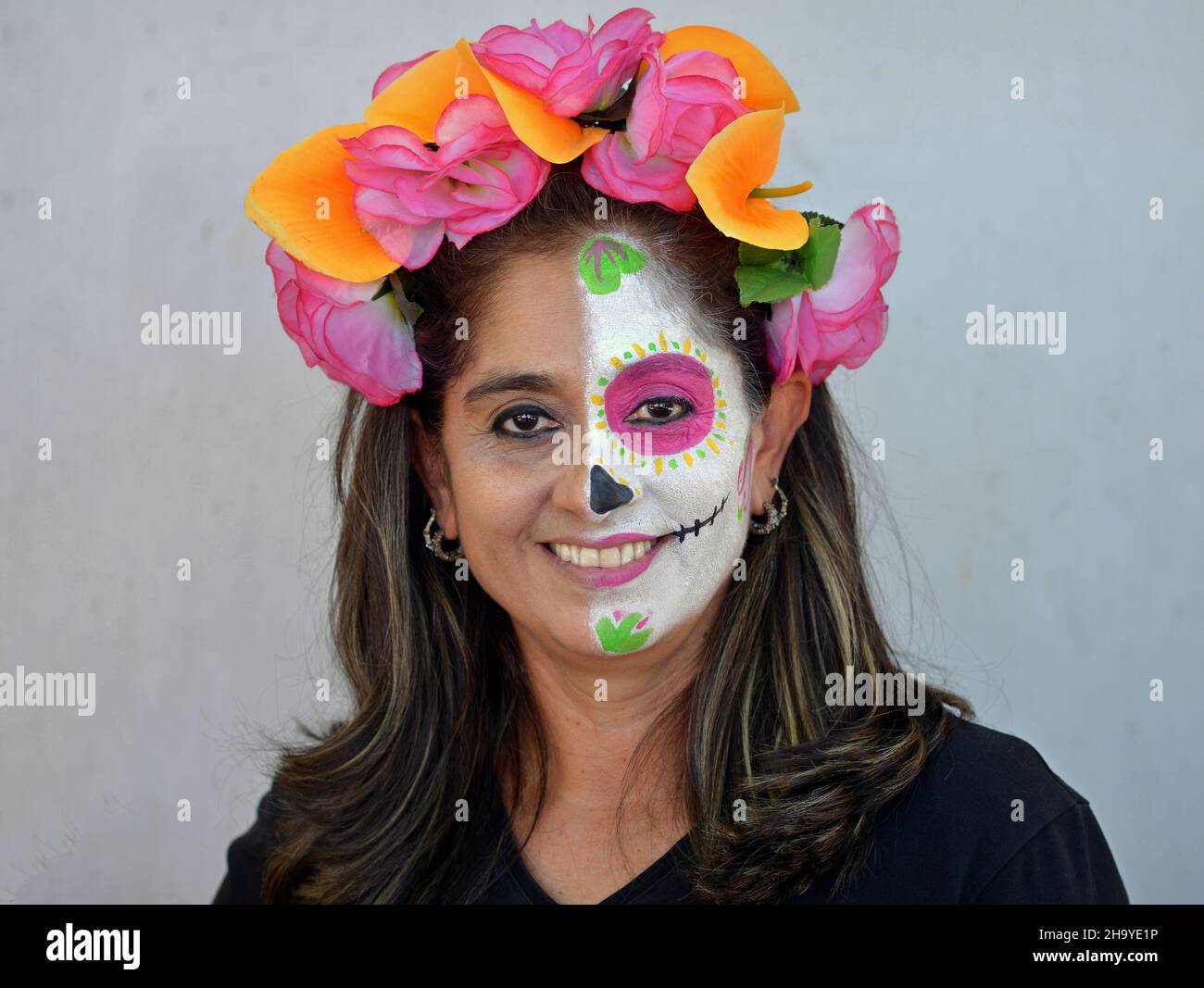 Mexikanische Frau mit bunten Blumen im Haar malt die linke Hälfte ihres Gesichts mit Catrina Face Make-up am Tag der Toten und lächelt den Betrachter an. Stockfoto