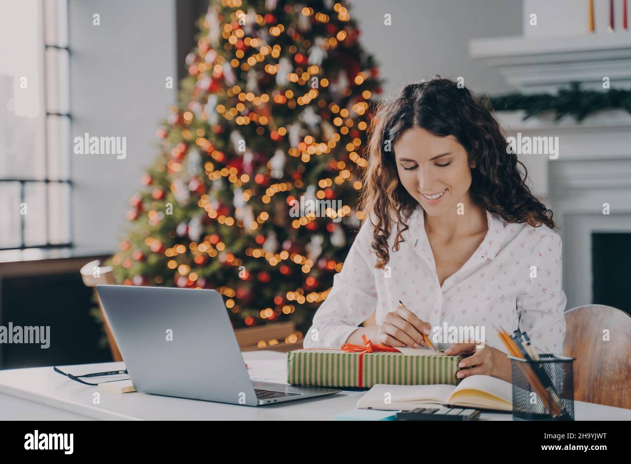 Lächelnde italienische Geschäftsfrau, die Weihnachtskarte schreibt und Geschenkbox für den Kunden verpackt Stockfoto