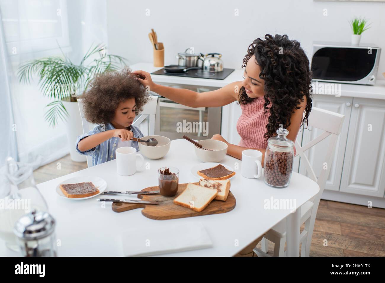 Lächelnde afroamerikanische Frau berührt das Kind und isst Getreide in der Nähe des Frühstücks zu Hause Stockfoto