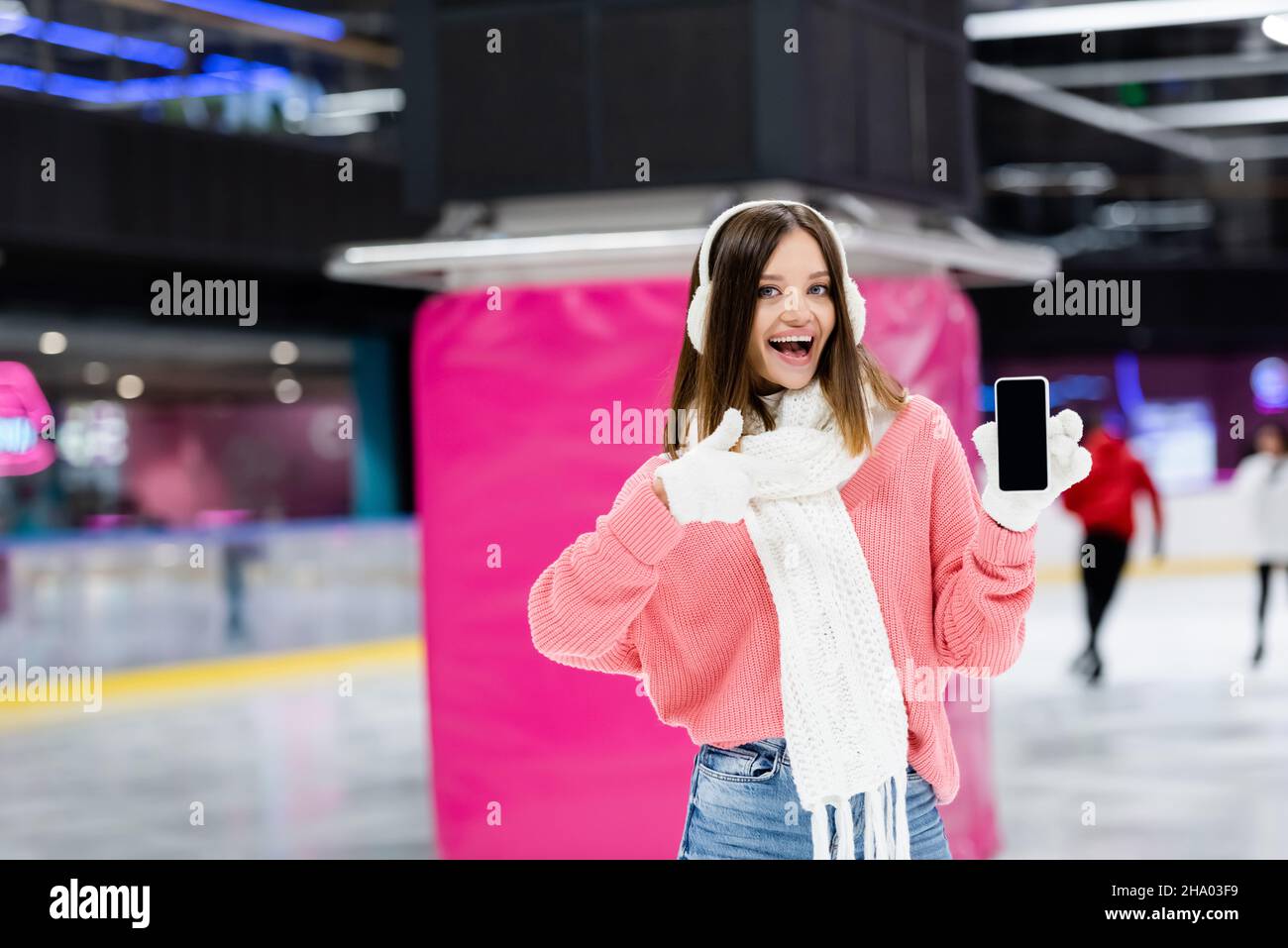 Staunende Frau in weißen Ohrenschützern, die das Smartphone mit leerem Bildschirm hält und den Daumen auf der Eisbahn zeigt Stockfoto