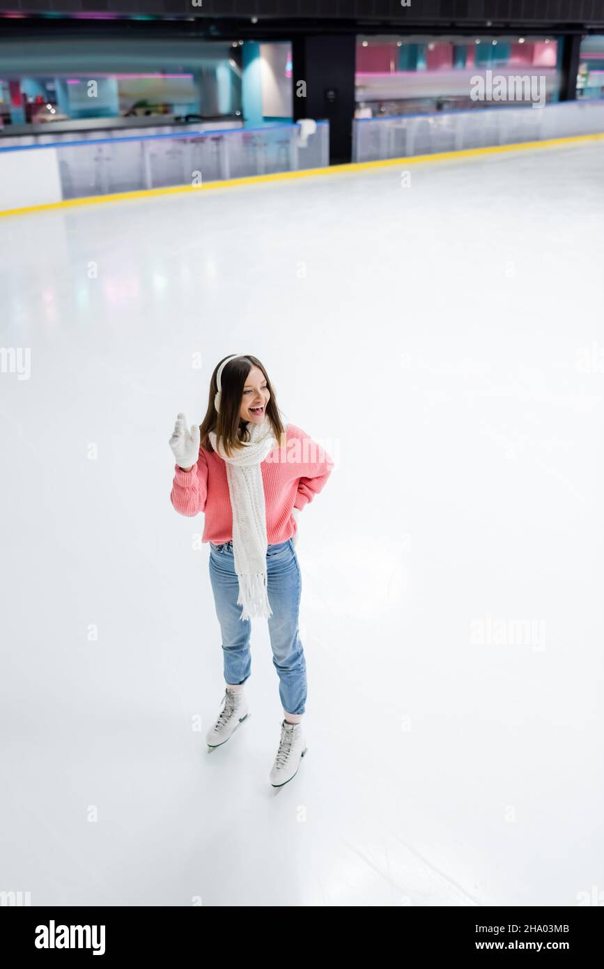 Die ganze Länge der glücklichen Frau posiert mit der Hand auf der Hüfte und winkt mit der Hand auf der Eisbahn Stockfoto