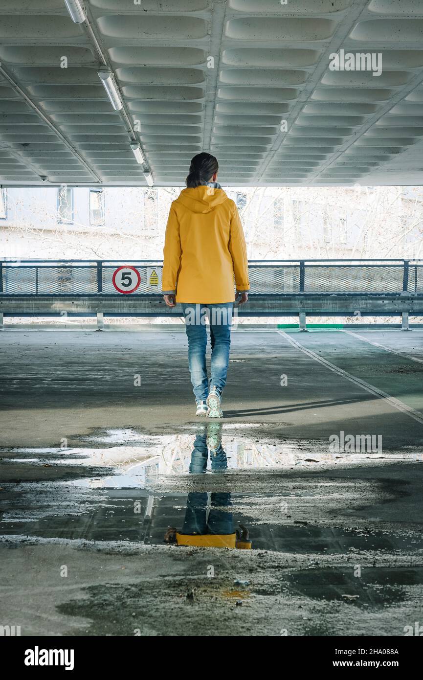 Rückansicht einer Frau, die in einem Betonparkplatz in einer gelben Jacke davonläuft. Stockfoto