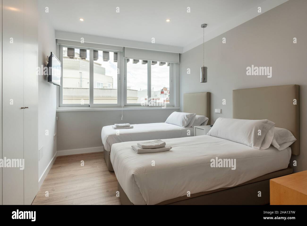 Schlafzimmer mit zwei separaten Betten mit Handtüchern und weißen Schränken,  wandfernseher und großem Fenster mit Blick in eine Ferienwohnung  Stockfotografie - Alamy