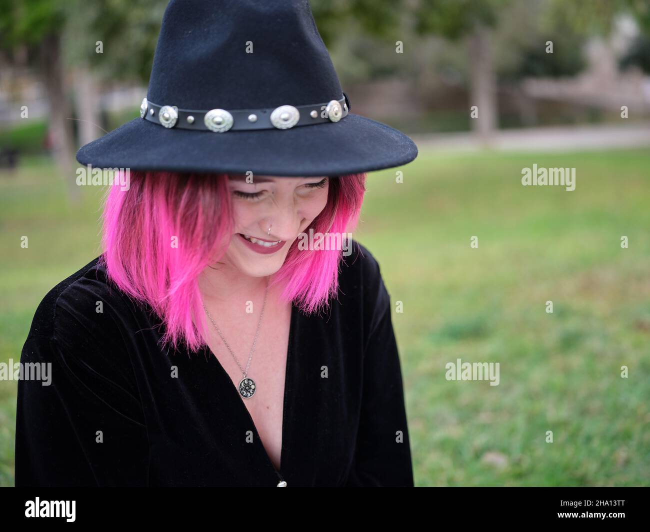Junge Frau mit rosa Haaren lächelt und schaut nach unten, trägt einen Hut und eine schwarze Bluse Stockfoto