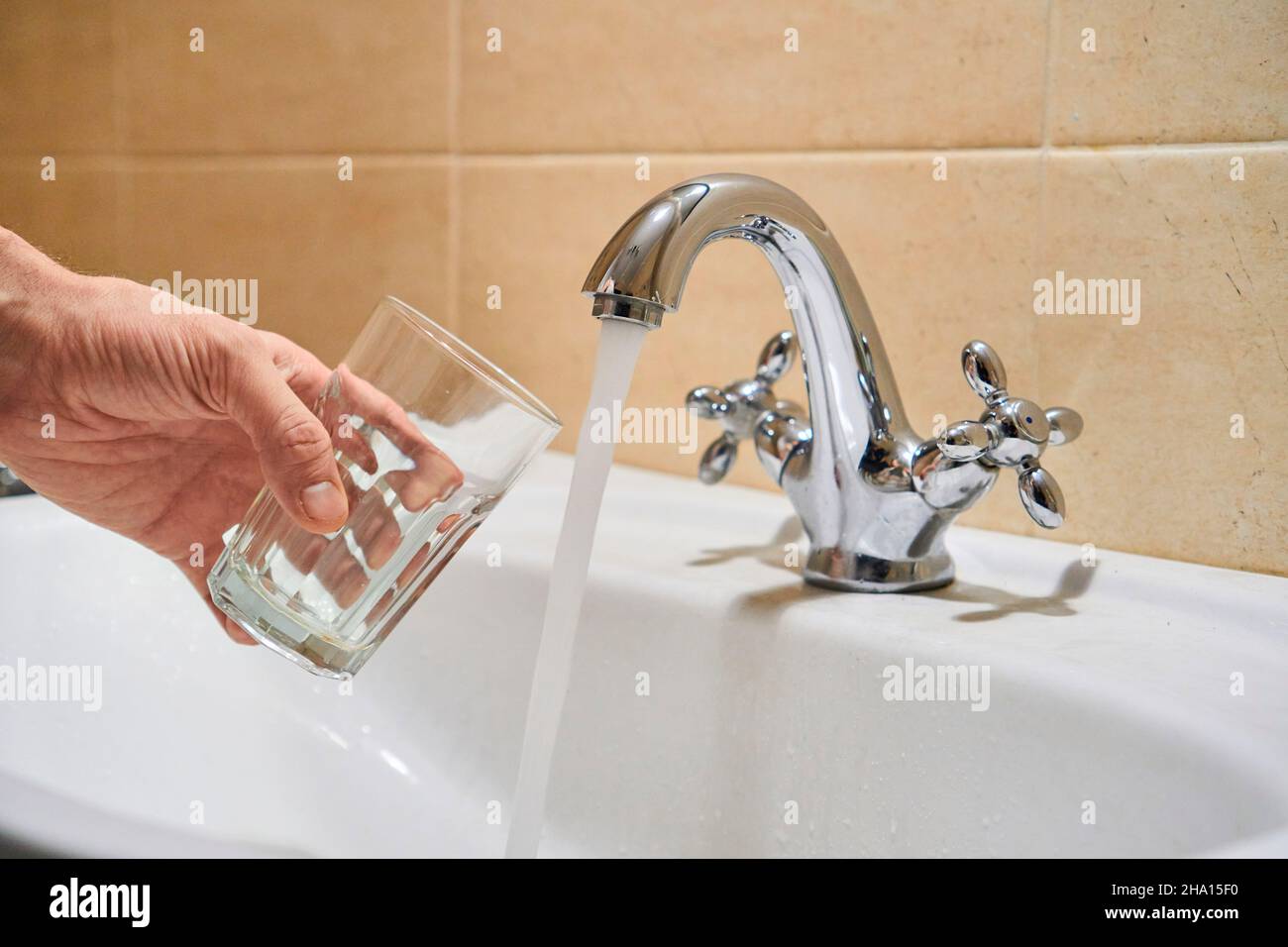 Mans Hand hält leeres Glas in der Nähe Badezimmer Edelstahl Säule Wasserhahn.  Erwägen Sie, fließendes Wasser aus dem Chrom-Wasserhahn zu gießen. Sicher zu  trinken oder nicht fit Stockfotografie - Alamy