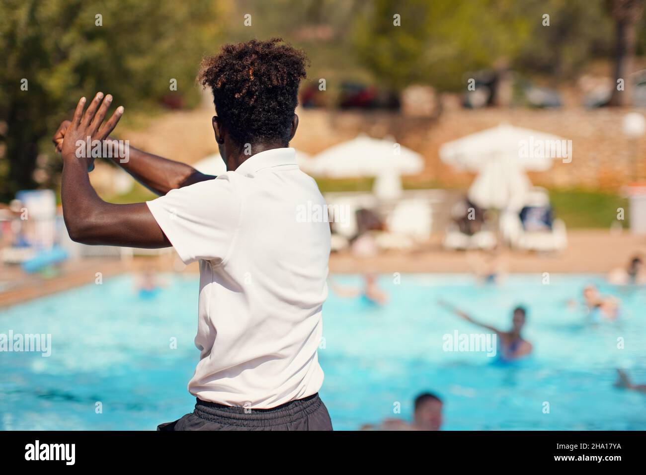 Der schwarze, sportliche Animator des Hotelresort trainiert vor dem Pool, Blick von der Seite hinter dem Hintergrund der verschwommenen Menschen - Wasseraerobic-Aktivitäten Stockfoto