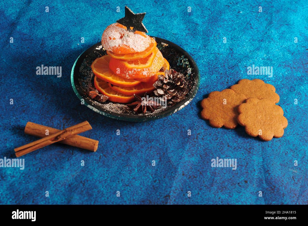 Aus orangefarbenen Scheiben gefertigte, winklige Ansicht eines winzigen Weihnachtsbaums. Blauer Backgroun mit Zimtstangen und Ingwerbrot. Stockfoto