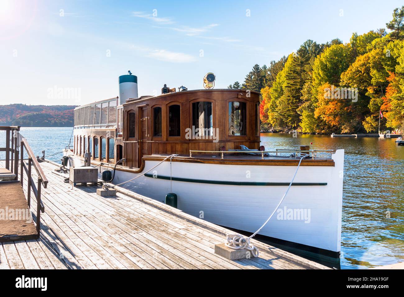 Altes Dampfschiff, das an einem sonnigen Tag an einem hölzernen Pier am See mit bunten Herbstbäumen im Hintergrund vertäut ist Stockfoto