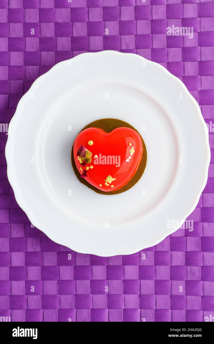 Ein Kuchen in Herzform. Ein herzförmiger Mousse-Cupcake auf einem Teller auf violettem Hintergrund. Valentinstag. Vertikales Foto Stockfoto