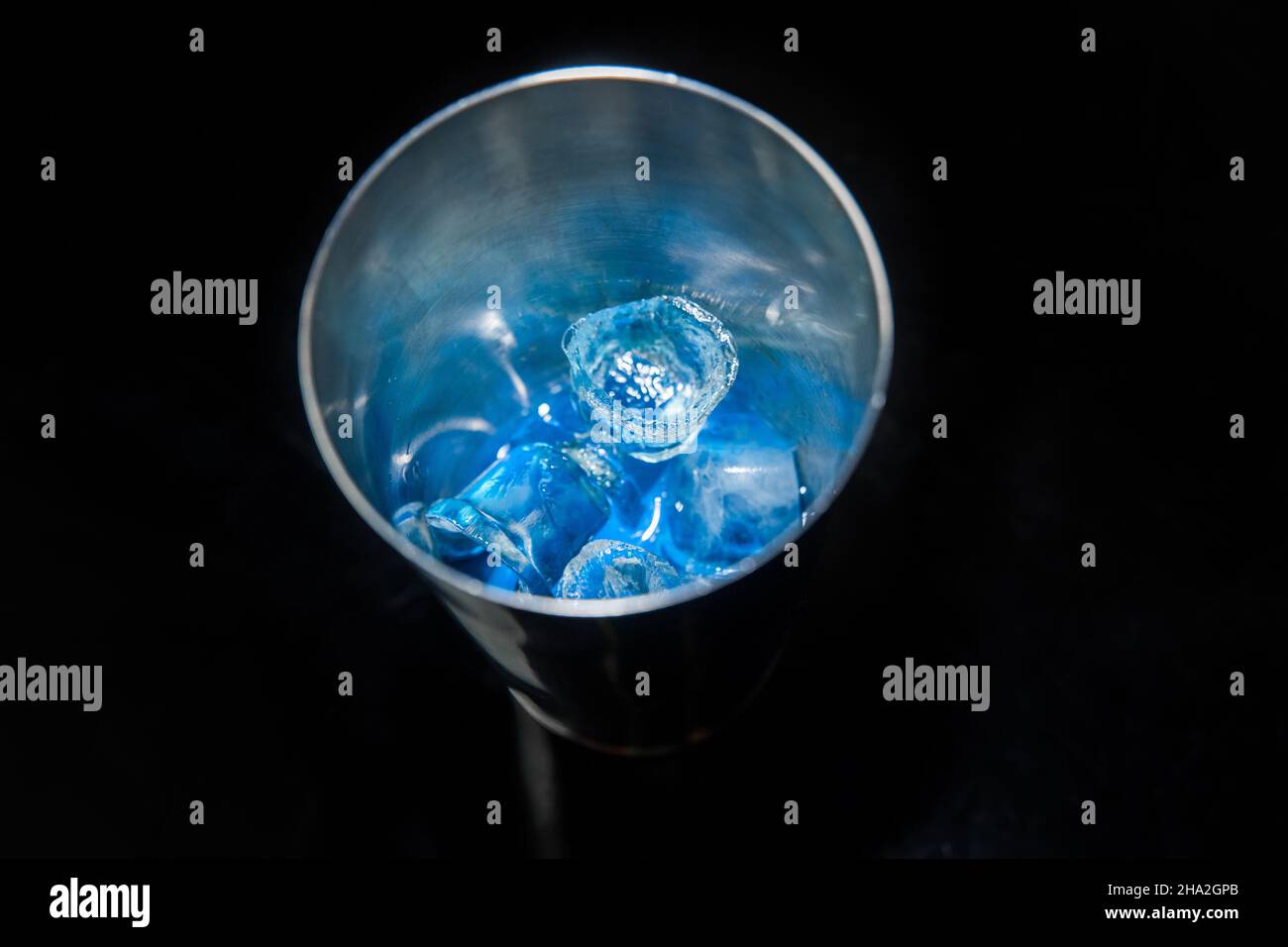 Blauer, kalter Sirup mit Eis in einem Werkzeug zum Mischen und Herstellen alkoholischer Cocktails Shaker auf schwarzem Hintergrund, Nahaufnahme. Stockfoto