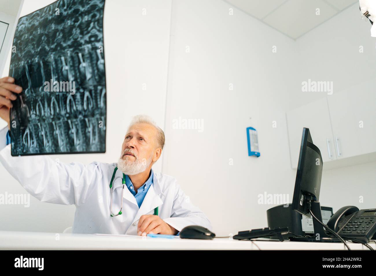 Niedriger Winkel des männlichen Arztes eines schweren älteren Erwachsenen, der das MRT-Wirbelsäulenbild des Patienten untersucht, der am Schreibtisch mit Computer im medizinischen Büroraum sitzt Stockfoto