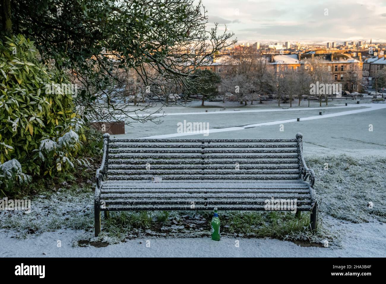 Eine unbesetzte Parkbank und eine leere Bierflasche auf dem schneebedeckten Boden an einem Wintertag im Queens Park, Glasgow. Stockfoto