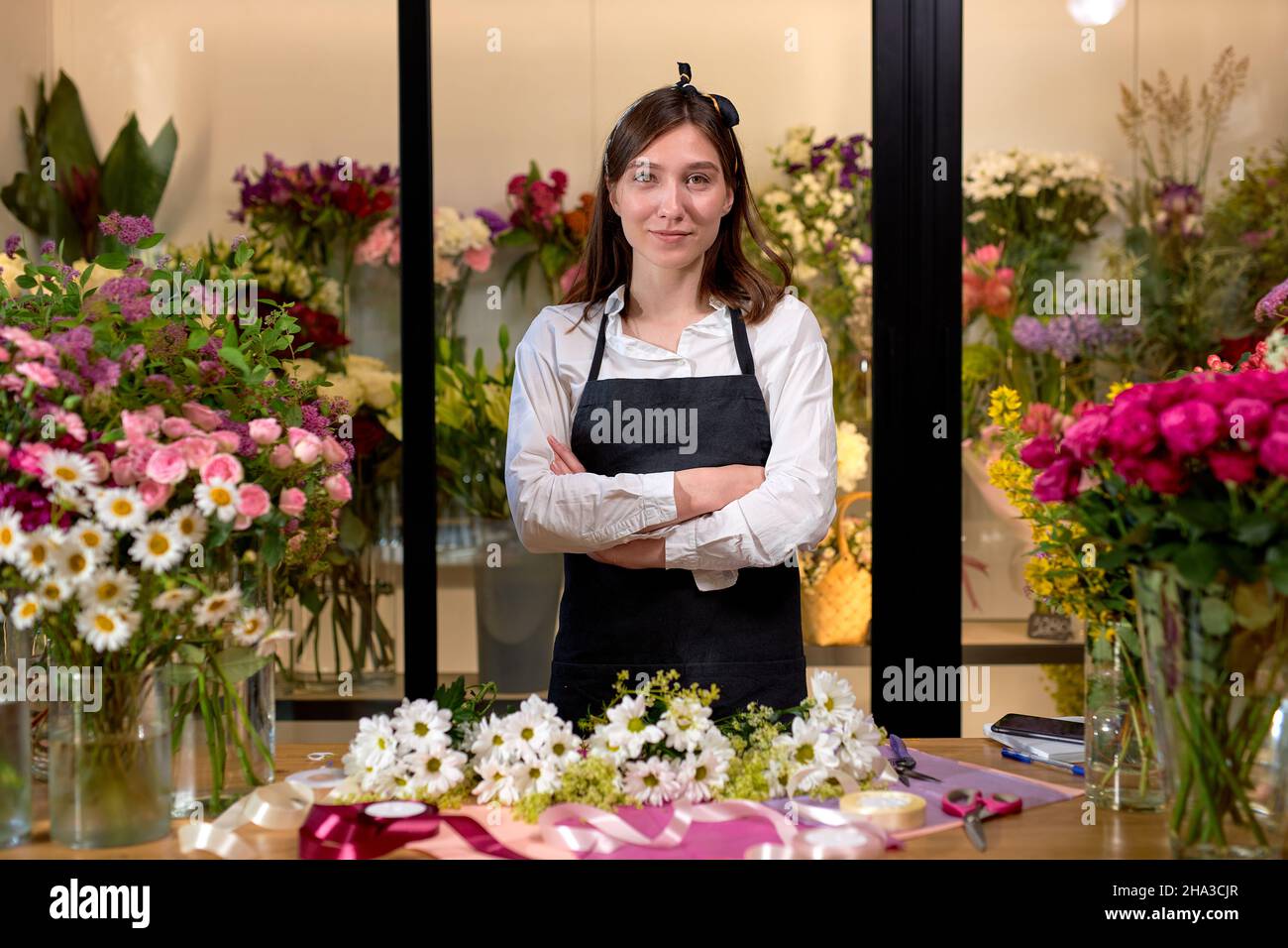 Junge lächelnde Floristen Shop Besitzer von Blumen umgeben, posiert mit Armen gefaltet. Schöne Dame lächelt, arbeitet im Blumenladen, schaut auf Kamera ha Stockfoto