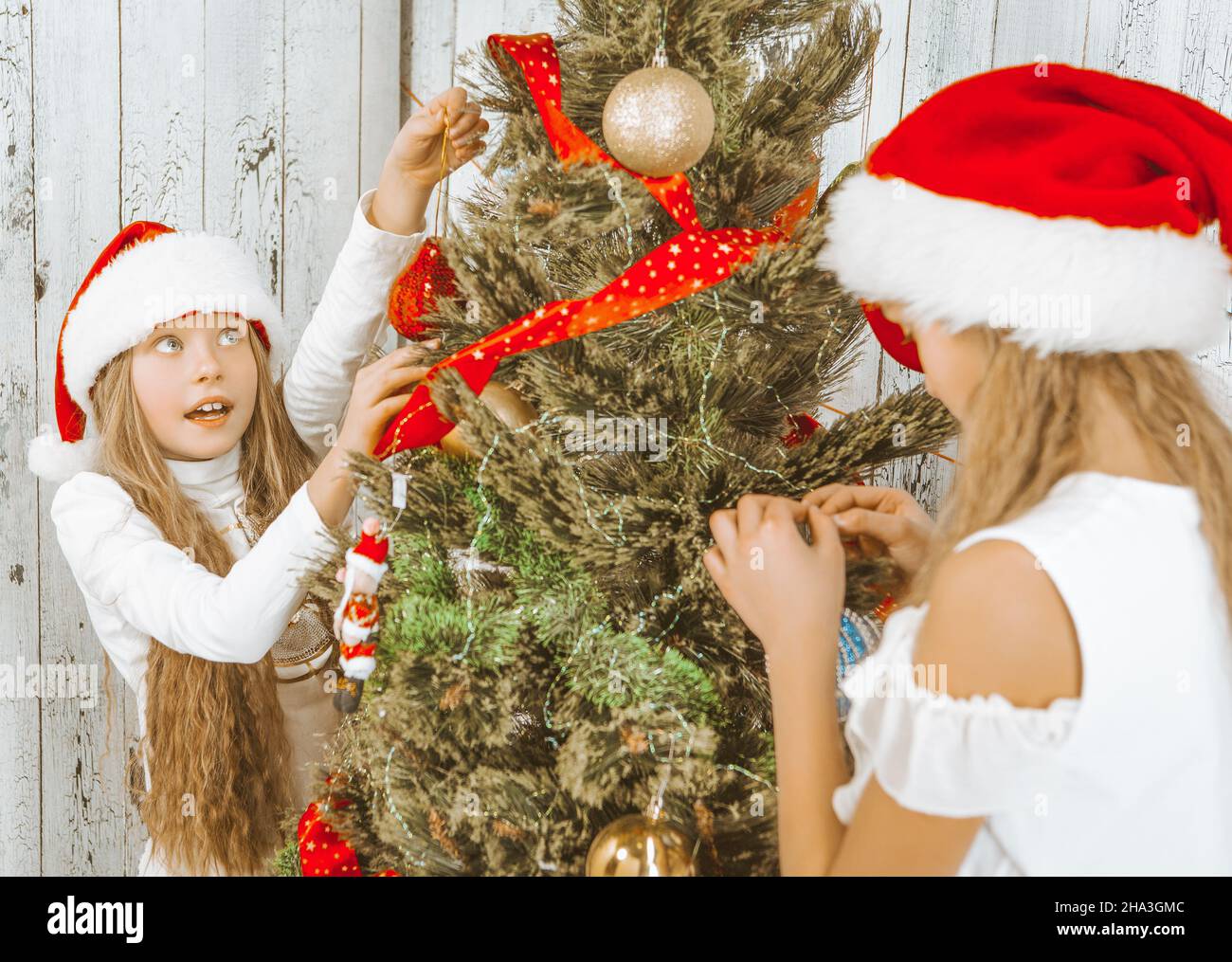 Weihnachtsmädchen schmücken den Weihnachtsbaum zusammen mit dekorativen Elementen. Zwei Schwestern hängen Dekorationen an einem Tannenbaum in ihrem Wohnzimmer zu Hause. Holzhintergrund. Nahaufnahme. Hochwertige Fotos Stockfoto