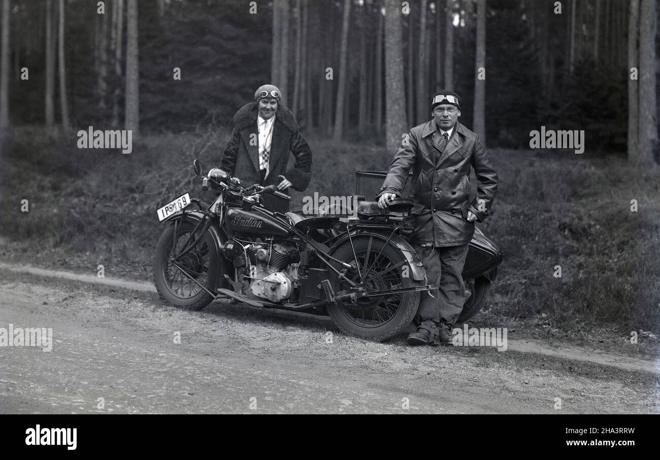 1930s, historisch, ein Gentleman und eine Dame, in Mänteln und Hüten mit Gesichtsgoogeln, stehen neben ihrem Motorrad, ein Indianer Scout, mit Seitenwagen, auf einem Schotterweg neben einem Wald, Frankreich. Der ikonische 'Indian Scout' war ein Motorrad, das von 1920 bis 1949 gebaut wurde und im Jahr WW11 eine gemeinsame Stätte war, als die 741, eine speziell gefertigte Version, vom Militär benutzt wurde. Stockfoto