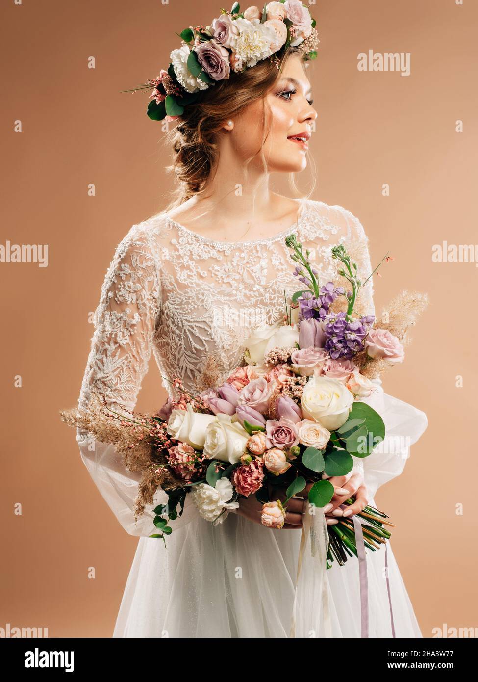 Wunderschöne lächelnde Braut im weißen Hochzeitskleid im Studio mit Blumenkranz und großem lila Bouquet. Hochzeit Floristen Trends. Brautmode. Stockfoto