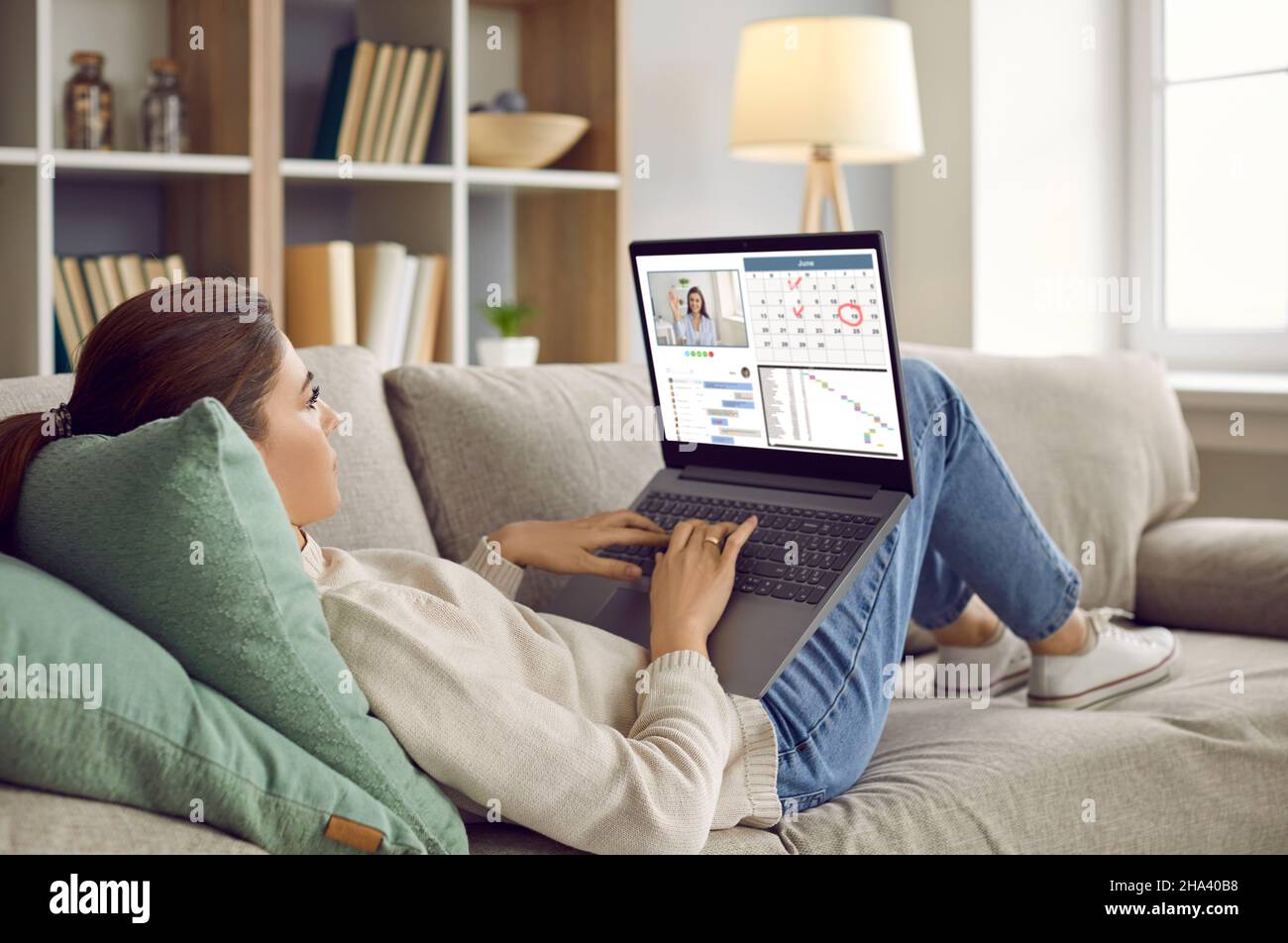 Frau, die zu Hause auf dem Sofa liegt, am Laptop arbeitet, den Kalender nutzt, im Arbeitsgespräch kommuniziert Stockfoto