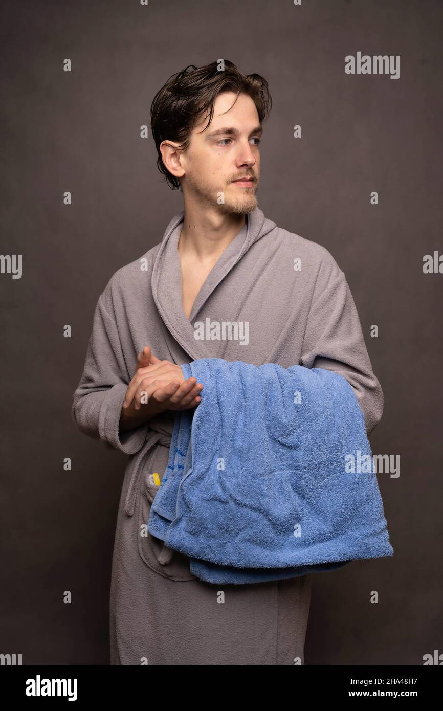 Junger, hübscher, großer, schlanker, weißer Mann mit braunen Haaren, das Handtuch in einem grauen Bademantel auf grauem Hintergrund hält Stockfoto