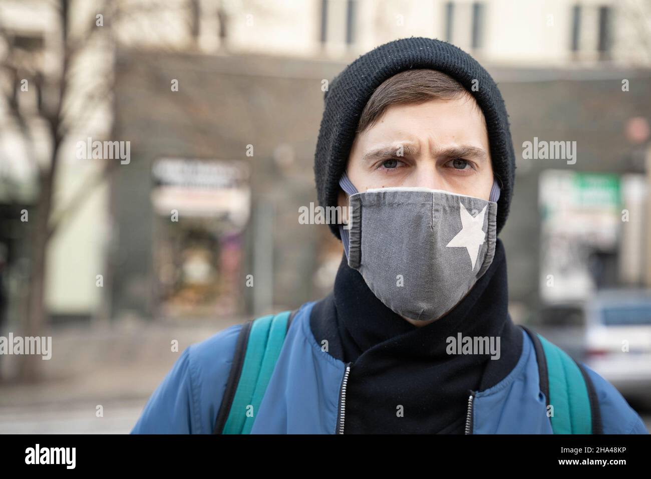 Der junge, hübsche weiße Mann mit braunen Haaren in blauer Jacke und schwarzem Winterhut runzelt die Stirn mit der Straßenszene im Hintergrund Stockfoto