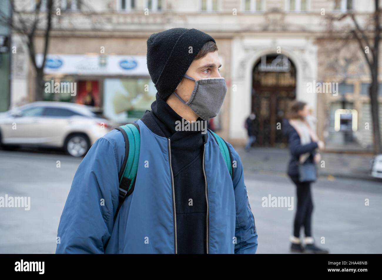 Junger hübscher weißer Mann mit braunem Haar in blauer Jacke mit schwarzem Winterhut schaut seitwärts mit der Straßenszene im Hintergrund Stockfoto