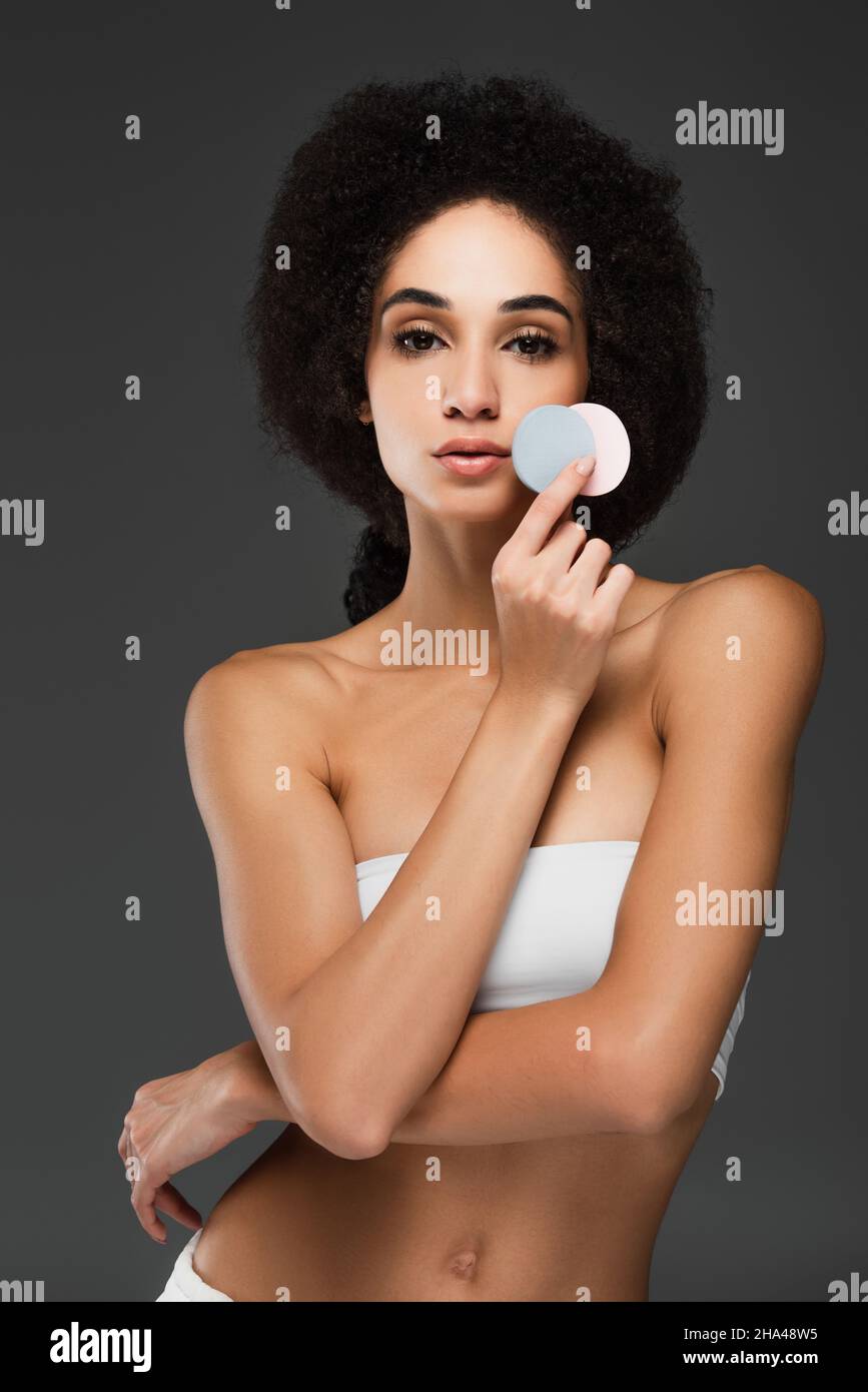 Schlanke afroamerikanische Frau mit perfektem Körper und Haut mit runden kosmetischen Schwämmen isoliert auf grau Stockfoto