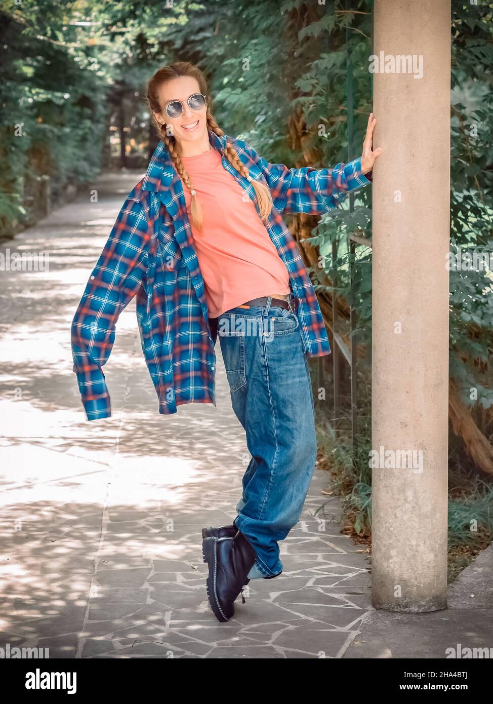 Ein Mädchen in einem karierten Hemd mit übermäßig langen Ärmeln und riesigen Jeans steht auf ihren Zehen im Park auf einer grünen Gasse Stockfoto