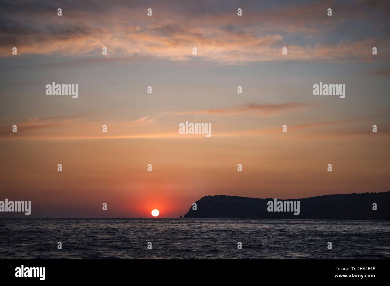 Dramatische Meereslandschaft über der schwarzen Meeresoberfläche während des Sonnenuntergangs, Silhouette der felsigen vulkanischen Klippe wird durch den warmen Sonnenuntergang beleuchtet. Stockfoto