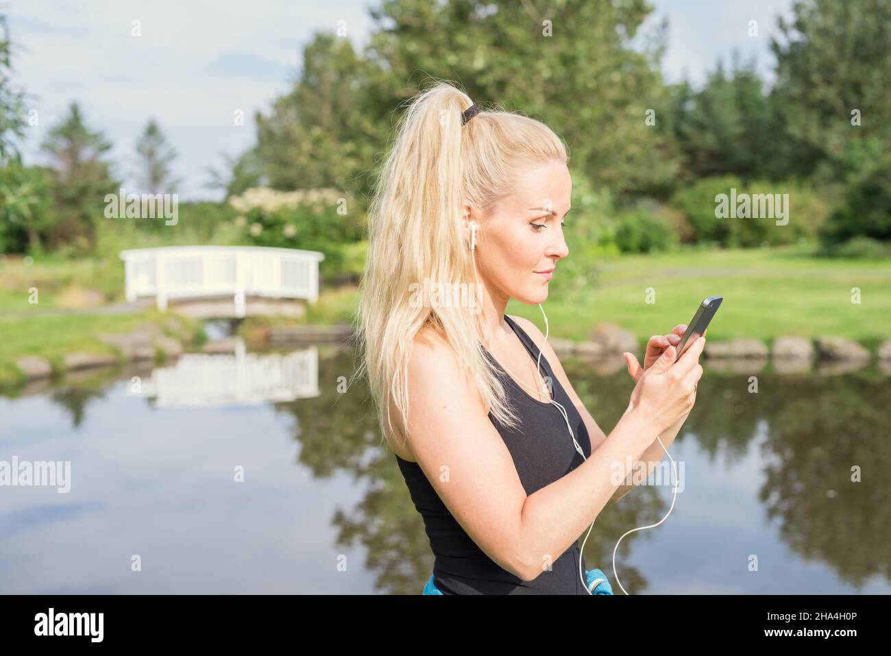 Seitenansicht einer selbstbewussten erwachsenen Sportlerin mit langem blondem Pferdeschwanz in Sportkleidung und Kopfhörern, die in der Nähe von Teichduerin auf dem Smartphone surfen Stockfoto