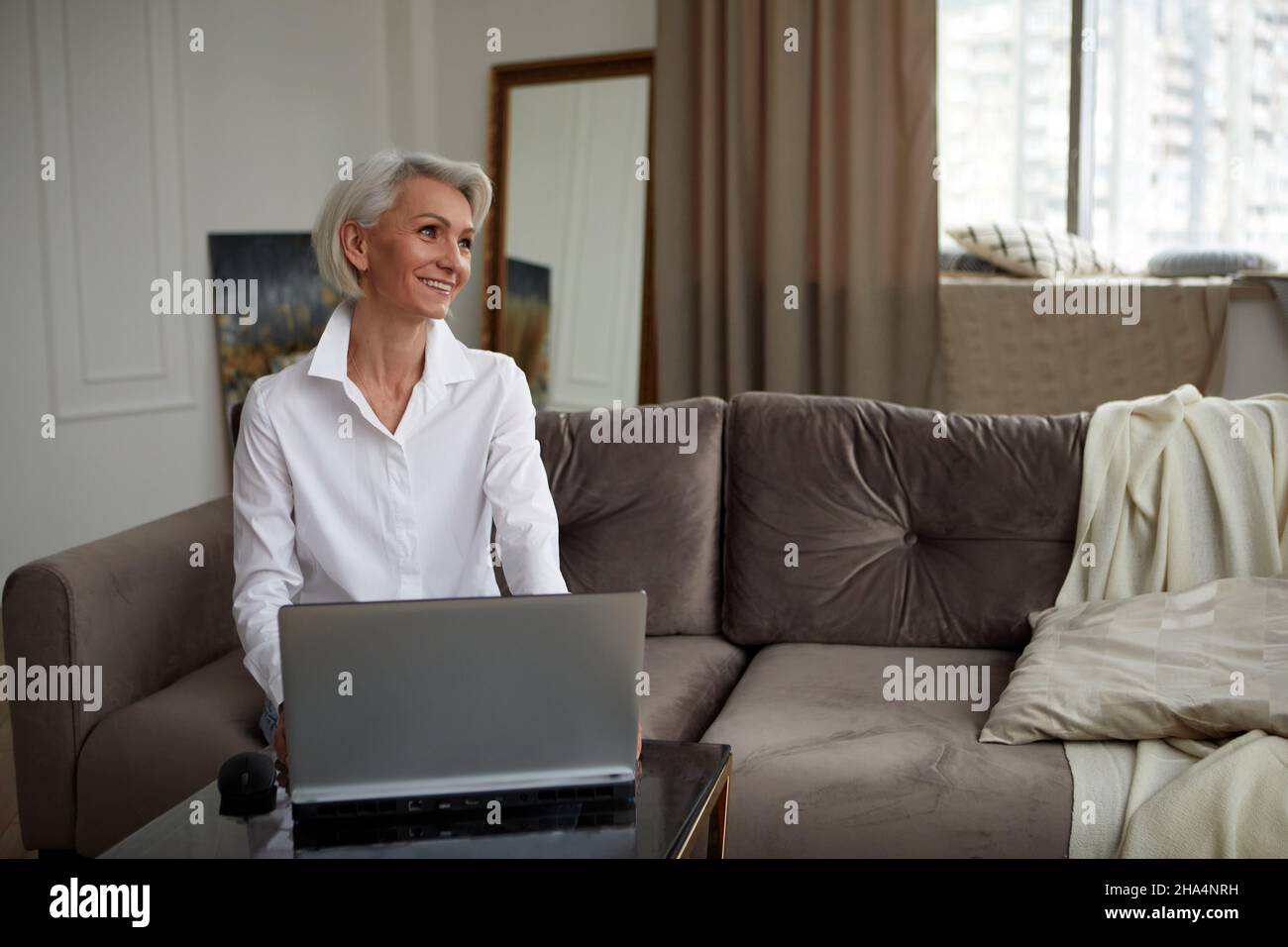 Glückliche 50s ältere ältere ältere Erwachsene Frau mittleren Alters mit Laptop mit Computer sitzen auf der Couch zu Hause. Lächelnde elegante ältere grauhaarige Dame spendin Stockfoto